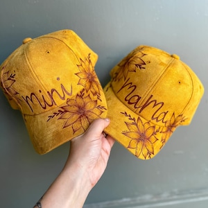 May include: Two yellow suede baseball caps with brown floral designs and the words "mini" and "mama" written on them.