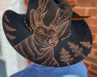 May include: A black felt cowboy hat with a brown deer design and a brown feather accent. The deer has antlers and is looking to the left. The hat has a brown leather band with a braided design.