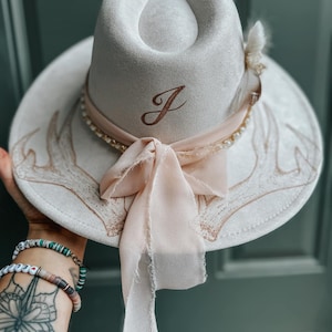 May include: A white felt fedora hat with a brown leather band and a pearl trim. The hat has a brown embroidered letter "J" on the front and a beige ribbon tied in a bow. The hat is decorated with a white antler design.