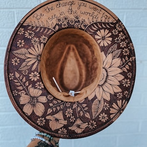 Puede incluir: Un sombrero marrón de ala ancha con un diseño floral quemado en la tela. El sombrero tiene una banda marrón alrededor de la corona y las palabras "Be the change you want to see in the world" están escritas en la banda.