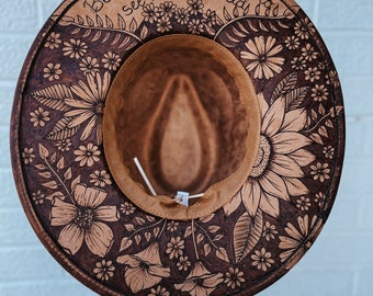 May include: A brown wide-brimmed hat with a floral design burned into the fabric. The hat has a brown band around the crown and the words "Be the change you want to see in the world" are written on the band.