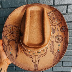 May include: A brown leather cowboy hat with a skull and dreamcatcher design. The hat has turquoise beads and feathers around the brim.