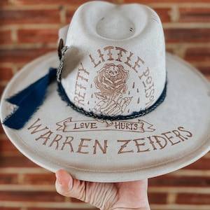 May include: A light gray fedora hat with the text "PRETTY LITTLE POISON" and "WARREN ZEIDERS" in brown. The hat features a rose with a skull graphic and a dark blue ribbon.