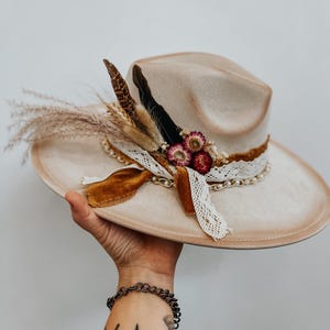 May include: A beige fedora hat with a wide brim, decorated with dried flowers, feathers, and a lace and chain band. The hat has a brown velvet ribbon. The hat is held up by a hand.