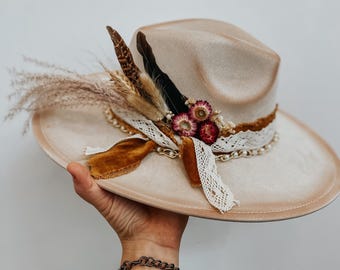 May include: A beige fedora hat with a wide brim, adorned with dried flowers, feathers, and a lace and chain band. The hat features a brown velvet ribbon and is held up by a hand.