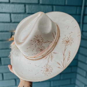 May include: A white felt fedora hat with a brown ribbon and a dandelion design. The hat has a wide brim and a tall crown.