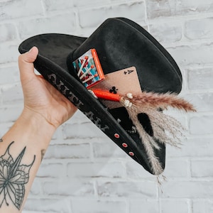 Puede incluir: Un sombrero vaquero negro con pedrería roja y la palabra "OUTLAW" bordada en el ala. El sombrero está decorado con una caja de cerillas, una carta de juego y un arreglo de flores secas.