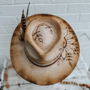 May include: A brown felt cowboy hat with a burnt design of a sunflower and leaves. The hat has a brown leather band and a feather accent.