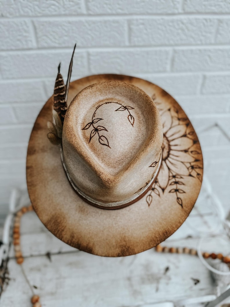 May include: A brown felt cowboy hat with a burnt design of a sunflower and leaves. The hat has a brown leather band and a feather accent.