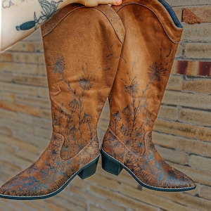 May include: A pair of brown suede knee-high boots with a floral design. The boots have a pointed toe and a low heel.