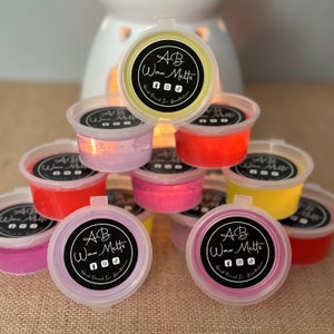 May include: A stack of small, clear plastic containers filled with colourful wax melts. The containers have black labels with the text "AB Wax Melts" and social media icons.