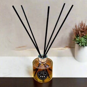 May include: A glass aromatherapy diffuser with a golden liquid and black reeds. The bottle has a silver collar and a black and white label with the text "AB Woo Multi". The diffuser is on a white and brown surface.