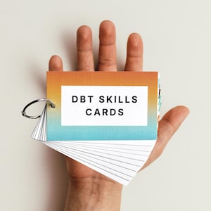May include: A hand holding a set of DBT Skills Cards. The cards are in a small, ring-bound booklet with a gradient cover that transitions from orange to blue.