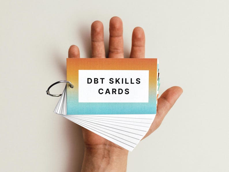 May include: A hand holding a set of DBT Skills Cards. The cards are in a small, ring-bound booklet with a gradient cover that transitions from orange to blue.