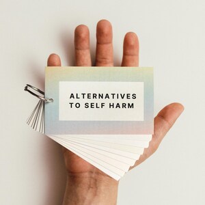 May include: A hand holding a small, rainbow-coloured booklet with the text "Alternatives to Self Harm" on the front cover.