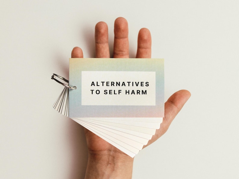 May include: A hand holding a small, rainbow-coloured booklet with the text "Alternatives to Self Harm" on the front cover.
