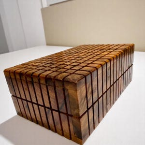 May include: A rectangular wooden box with a grid-like pattern carved into the top and sides. The box is made of a dark brown wood and has a hinged lid. The box is sitting on a white surface.