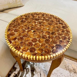 May include: A round side table with a mosaic-style wooden top. The tabletop has a geometric pattern in various wood tones. The edge is trimmed with a light-coloured border. The table has three curved legs.