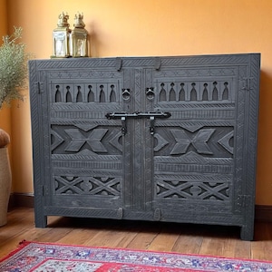 Può includere: Un mobile in legno grigio scuro, intagliato in modo intricato, con due ante. Il mobile presenta motivi geometrici e stilizzati, un chiavistello in metallo e maniglie ad anello. Due lanterne decorative sopra.
