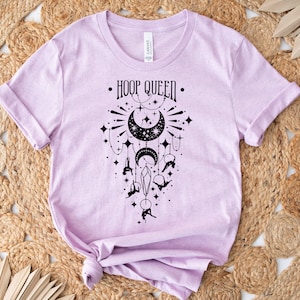 Könnte beinhalten: Ein lavendelfarbenes T-Shirt mit einem schwarzen Grafikdesign eines Halbmondes mit Sternen und einer Hula-Hoop-Tänzerin. Der Text "HOOP QUEEN" ist auf dem Shirt gedruckt.