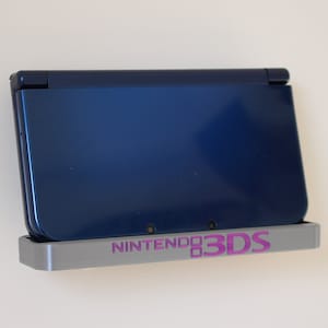 Puede incluir: Un soporte de pared para una consola de juegos portátil Nintendo 3DS. El soporte está hecho de metal plateado y tiene el logotipo de Nintendo 3DS impreso en rosa. Una Nintendo 3DS azul está en el soporte.