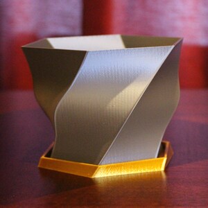 Puede incluir: Un macetero gris geométrico con una base hexagonal dorada. El macetero tiene un diseño único y retorcido.