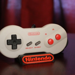 Puede incluir: Un mando de control gris de Nintendo Entertainment System con botones rojos y un logotipo de Nintendo negro en un soporte.