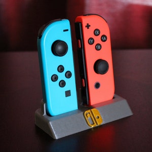 May include: Two Nintendo Switch Joy-Con controllers, one blue and one red, are displayed in a grey stand with a gold Nintendo logo.