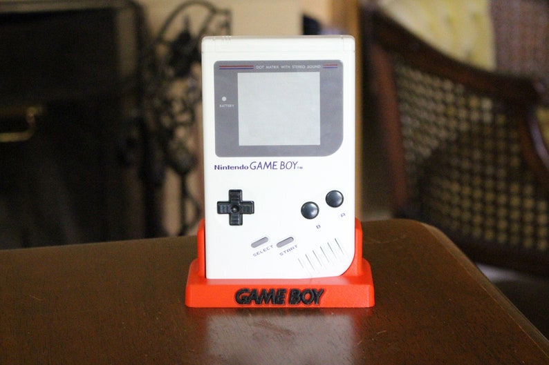 May include: A white Nintendo Game Boy handheld video game console with a gray screen and black buttons. The console is sitting on a red stand with the words "GAME BOY" printed on the front.