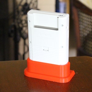 May include: A white Game Boy handheld console with a red plastic stand. The console is sitting on a wooden surface.