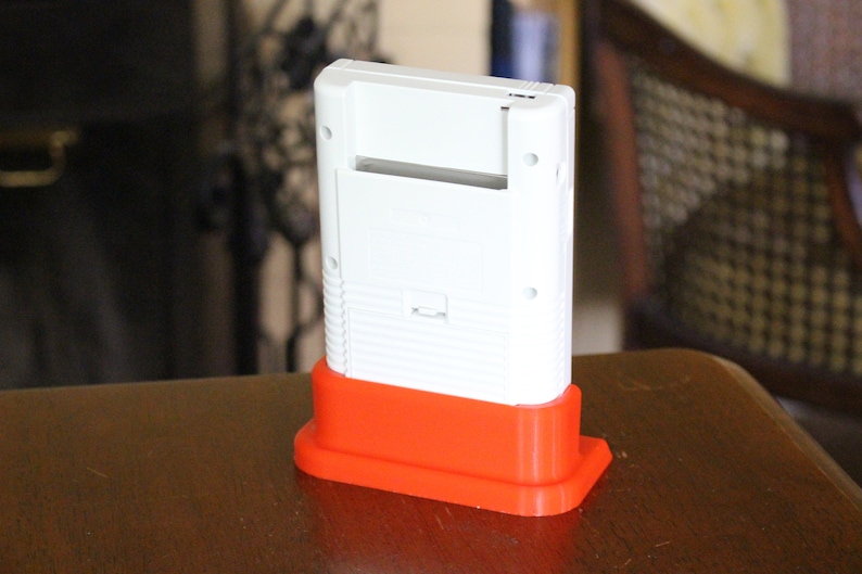 May include: A white Game Boy handheld console with a red plastic stand. The console is sitting on a wooden surface.