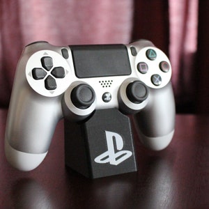 Pode incluir: Um comando PlayStation 4 prateado é exibido em um suporte preto com o logotipo do PlayStation. O comando tem botões pretos e um touchpad preto.