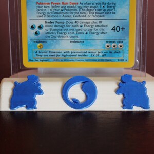May include: A white plastic card holder with a blue 3D printed Blastoise Pokemon card. The card holder has a round indentation for the card and two smaller indentations on either side. The card holder is sitting on a dark brown surface.