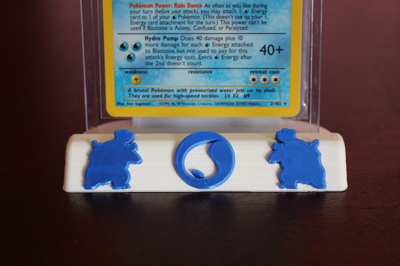 May include: A white plastic card holder with a blue 3D printed Blastoise Pokemon card. The card holder has a round indentation for the card and two smaller indentations on either side. The card holder is sitting on a dark brown surface.