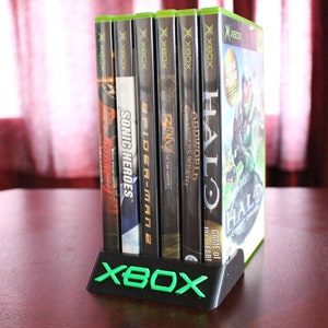 May include: A black plastic stand with the word "XBOX" in green letters holds a stack of six video game cases. The cases are all for the Xbox console and feature various game titles, including "Halo", "Oddworld", "Rynx", "Spider-Man 2", and "Sonic Heroes".