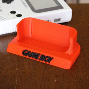 May include: A bright orange plastic stand with the words "GAME BOY" printed in black. The stand is designed to hold a Game Boy handheld gaming device.
