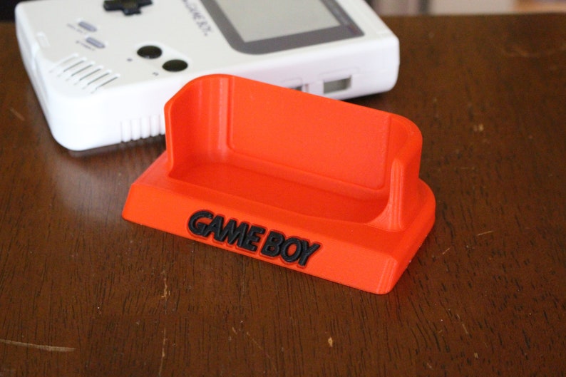 May include: A bright orange plastic stand with the words "GAME BOY" printed in black. The stand is designed to hold a Game Boy handheld gaming device.
