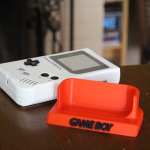 May include: A white Game Boy handheld video game console with a black and gray screen and a red plastic stand with the words "GAME BOY" printed on it.