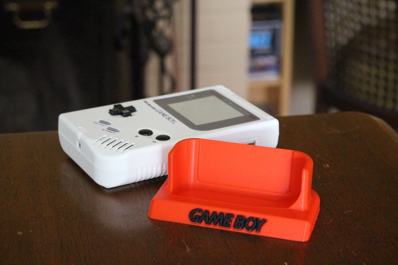 May include: A white Game Boy handheld video game console with a black and gray screen and a red plastic stand with the words "GAME BOY" printed on it.