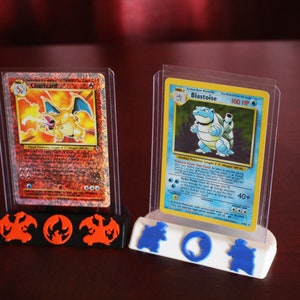 May include: Two Pokemon trading cards, Charizard and Blastoise, are displayed in clear plastic cases on a 3D printed stand. The stand has a red and black base with a Charizard silhouette and a blue and white base with a Blastoise silhouette.