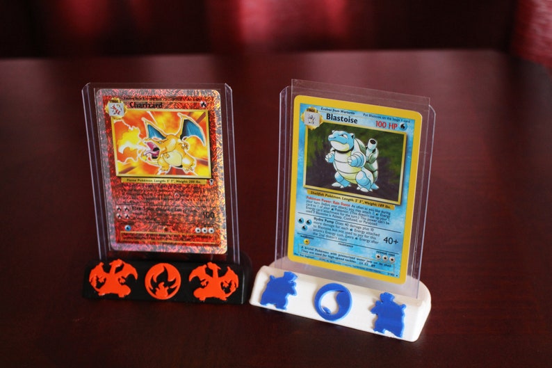May include: Two Pokemon trading cards, Charizard and Blastoise, are displayed in clear plastic cases on a 3D printed stand. The stand has a red and black base with a Charizard silhouette and a blue and white base with a Blastoise silhouette.