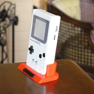 May include: A white Game Boy handheld video game console on a bright orange stand. The stand has the word "Gameboy" printed on it in black.