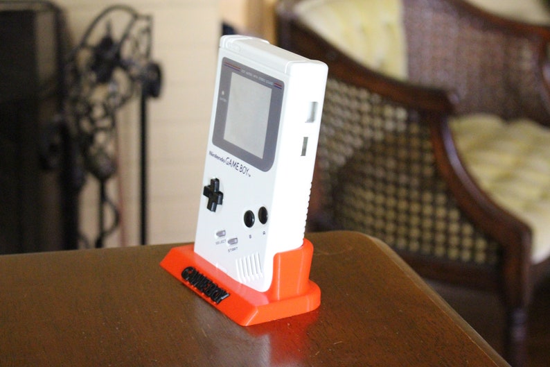 May include: A white Game Boy handheld video game console on a bright orange stand. The stand has the word "Gameboy" printed on it in black.