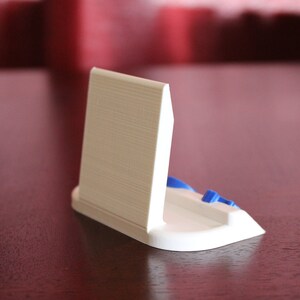 May include: White plastic phone stand with a blue accent. The stand is designed to hold a phone upright.