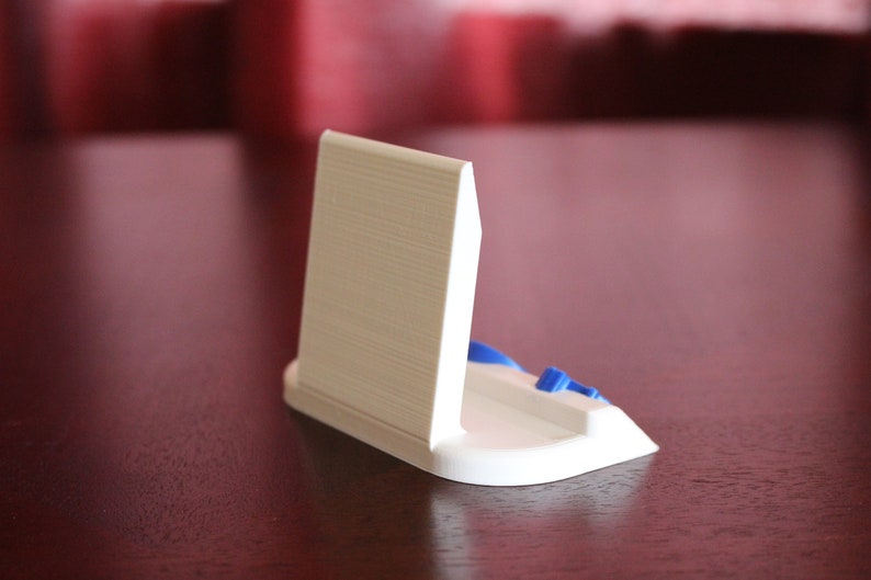 May include: White plastic phone stand with a blue accent. The stand is designed to hold a phone upright.