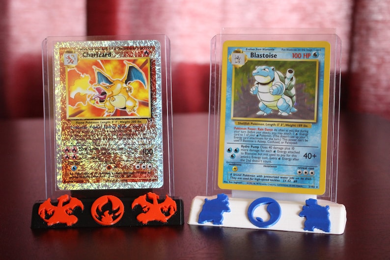 May include: Two Pokemon cards, Charizard and Blastoise, are displayed in clear plastic cases on a black and orange display stand. The stand has a dragon design on the left and right sides and a fire symbol in the center. The stand has a blue and white design on the right side with a water symbol in the center.