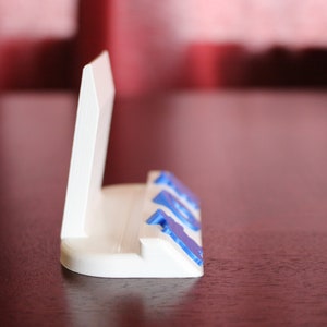 May include: White plastic phone stand with blue accents. The stand has a raised back to hold a phone upright.