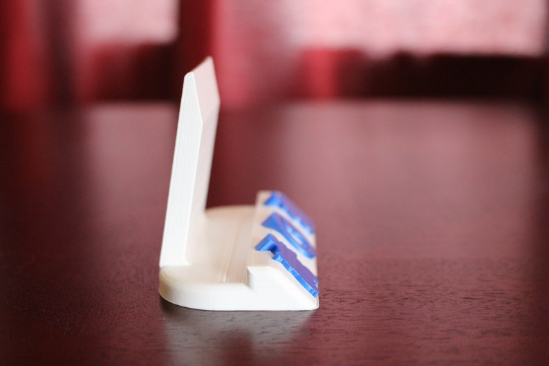 May include: White plastic phone stand with blue accents. The stand has a raised back to hold a phone upright.