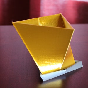 May include: A gold-colored triangular-shaped planter with a white base. The planter is made of a textured material and has a smooth, shiny finish.