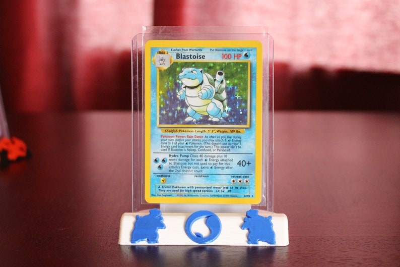 May include: A clear plastic case with a blue and white Pokemon card inside. The card features a blue and white Blastoise Pokemon with the text "Blastoise 100 HP" and "Shellfish Pokemon Length: 6'7" Weight: 189 lbs."